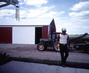 A guy using a white cane to navigate outside with a barn and tractor in the background.